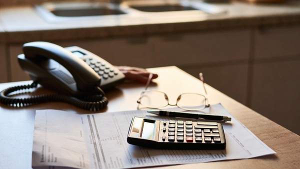 A paper bill on a kitchen table with a calculator, a pen and a corded telephone