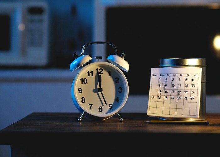 A twin-bell alarm clock and a small paper calendar on a kitchen table at dusk, a blank microwave display behind them
