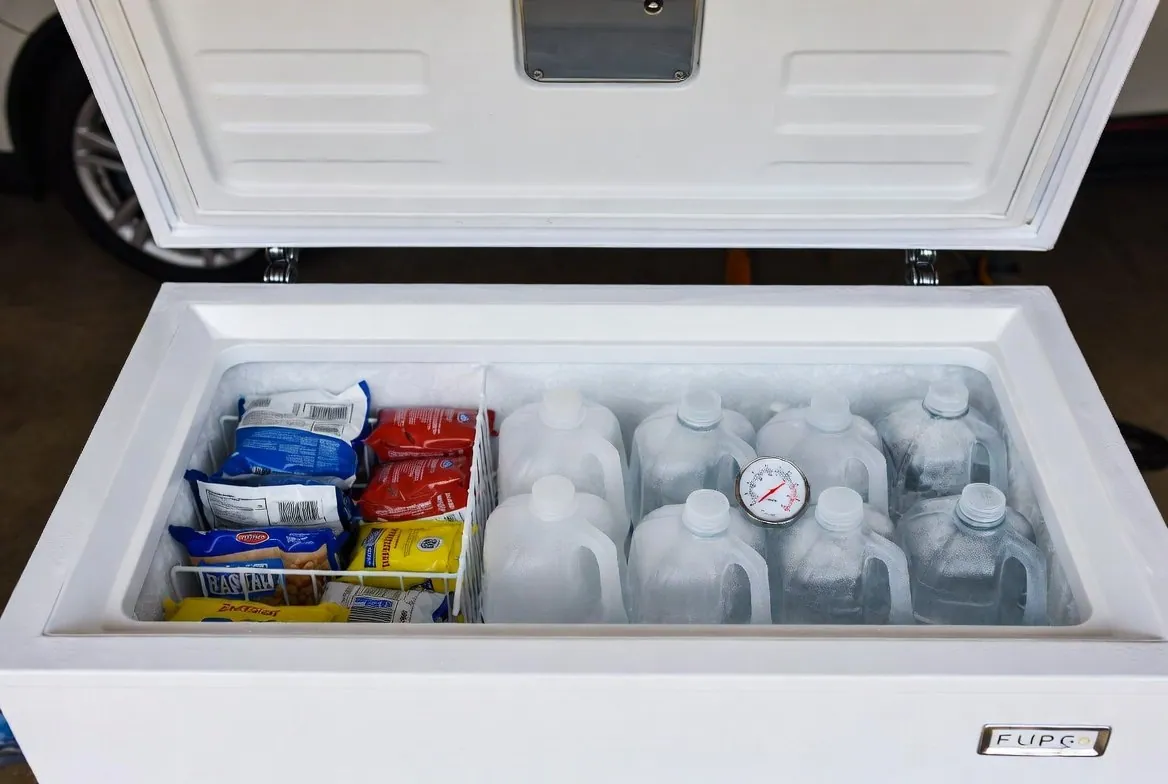 A chest freezer with its lid open, food packages in a basket on one side and rows of frozen one-gallon water jugs on the other, a dial thermometer resting on the jugs