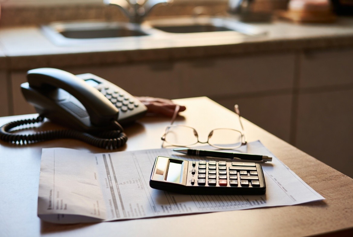 A paper utility bill spread open on a kitchen table with a pocket calculator on top of it, a pen, reading glasses and a corded telephone within reach