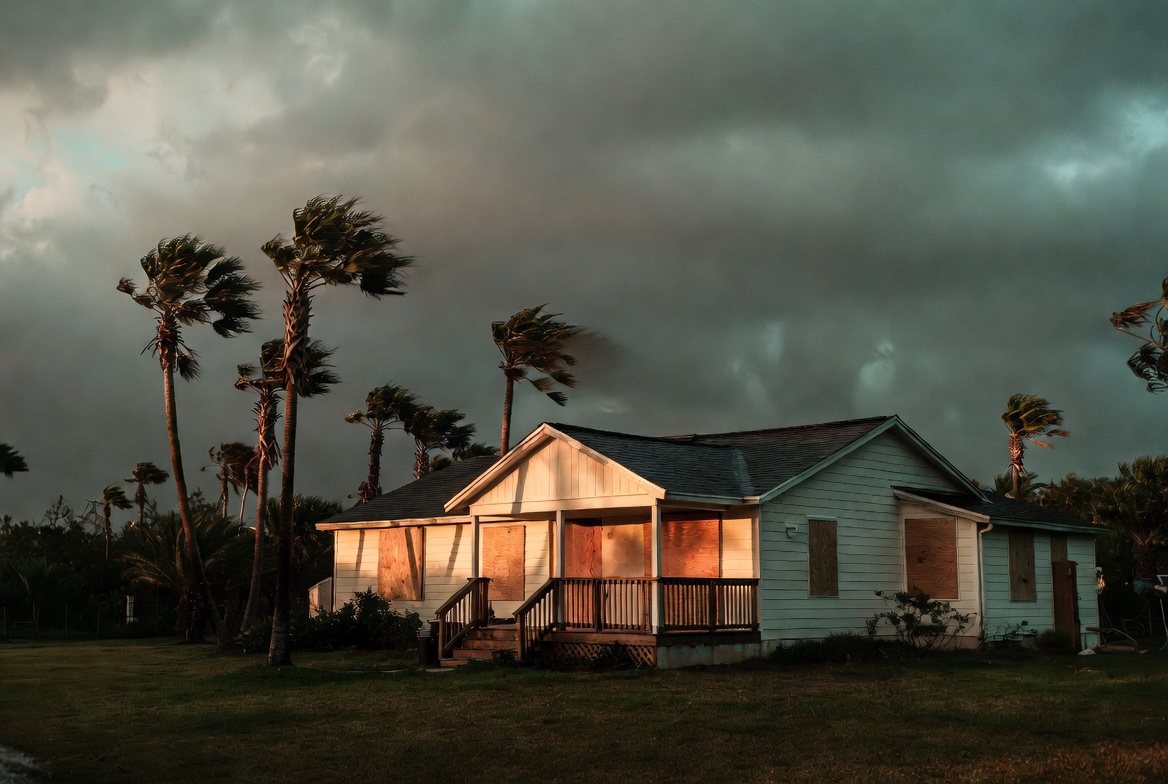 A single-storey Florida house with plywood screwed over every window, palms bending in rising wind under a heavy storm sky at dusk