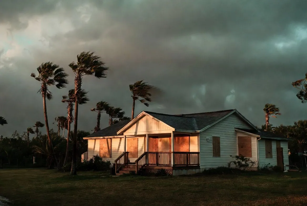 A single-story Florida house with plywood screwed over every window, palms bending in rising wind under a heavy storm sky at dusk