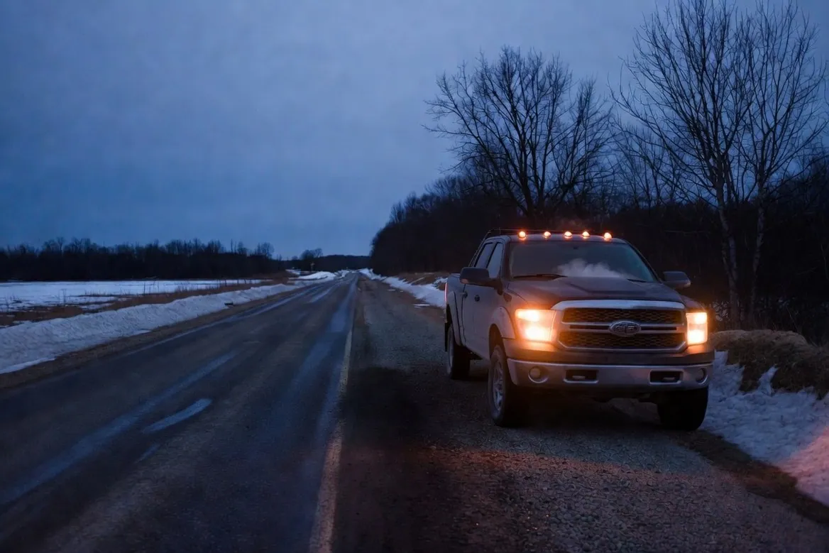 A pickup stopped on the gravel shoulder of an empty rural road at winter dusk, headlights and cab lights lit, a plume of steam rising from the hood, snow in the ditches and no other vehicle anywhere in sight