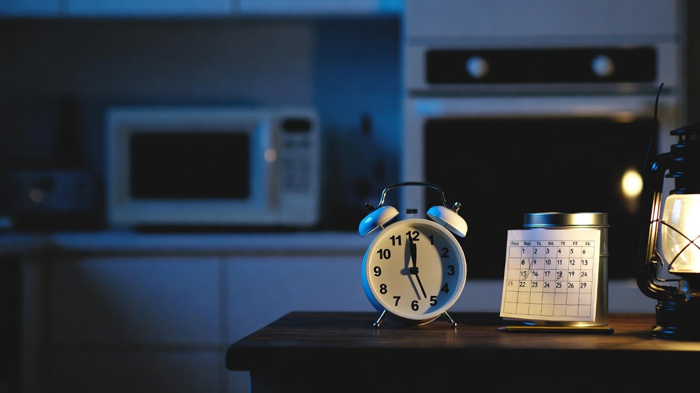 A twin-bell alarm clock and a small paper calendar standing on a kitchen table at dusk, with a microwave and a wall oven behind them showing completely blank displays