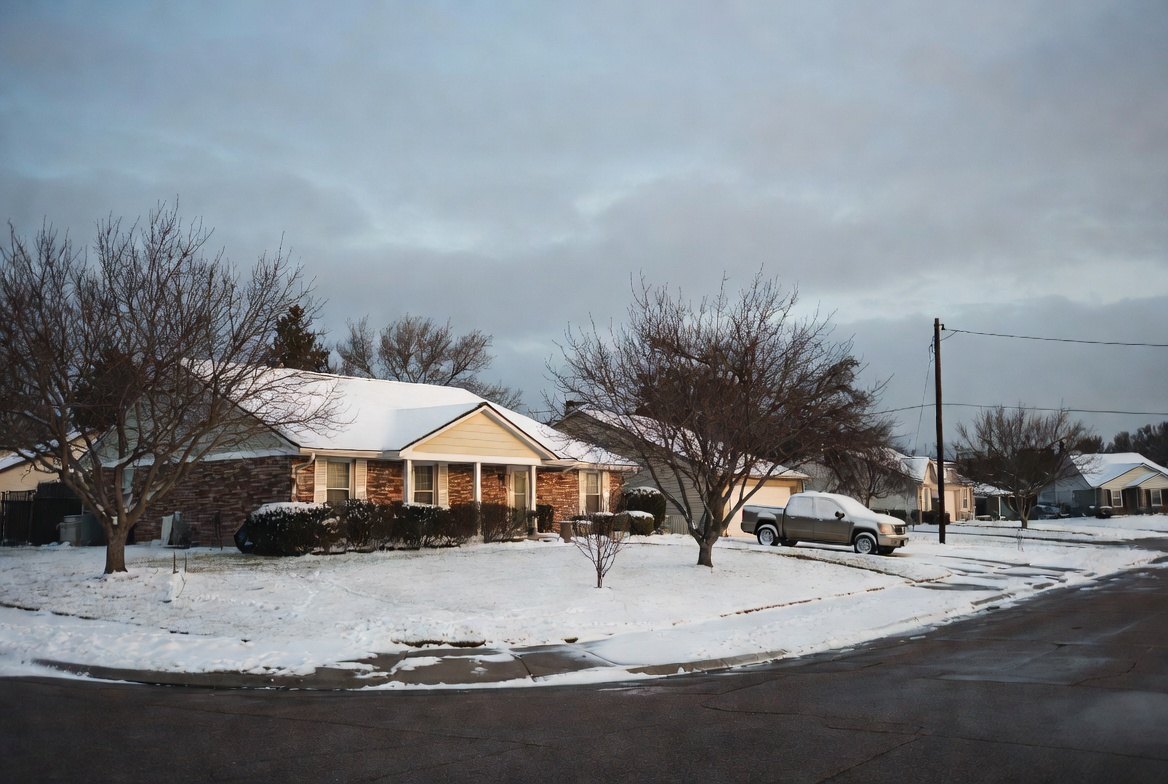 A snow-covered street of single-storey Texas brick houses under a heavy grey sky, bare trees and overhead power lines, one pickup truck in a driveway and no one outside