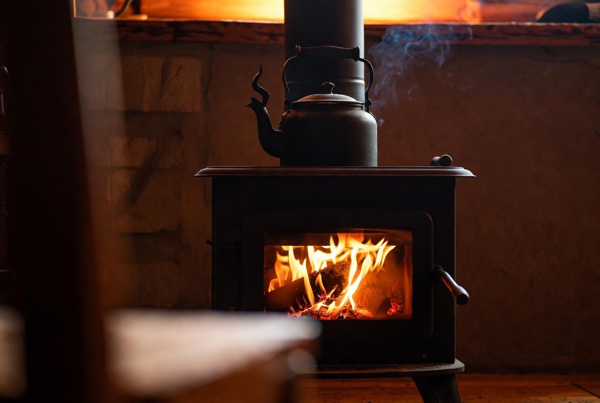 A cast-iron wood stove burning brightly in a dim room with a kettle steaming on top of it, stone wall behind and a doorframe in shadow to the left