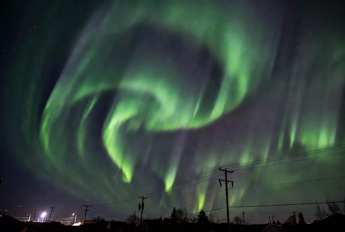 A green and violet aurora filling the night sky above an ordinary residential street with utility poles and power lines in silhouette below