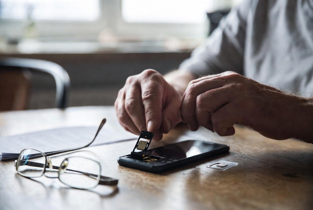 An older person’s hands at a kitchen table easing the SIM tray out of a plain black phone, with a new SIM card in its carrier on the table and reading glasses beside it