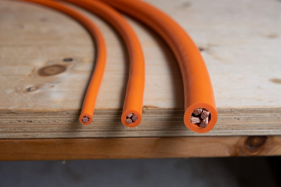 Three orange cables of increasing thickness laid side by side on a plywood bench, each cut square so the copper conductor inside is visible, the thickest carrying several times the metal of the thinnest