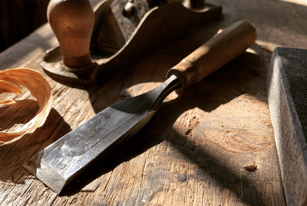 A bench chisel lying on a workbench with its bevel catching the light, a hand plane behind it, a curl of pine shaving to one side and a sharpening stone at the right