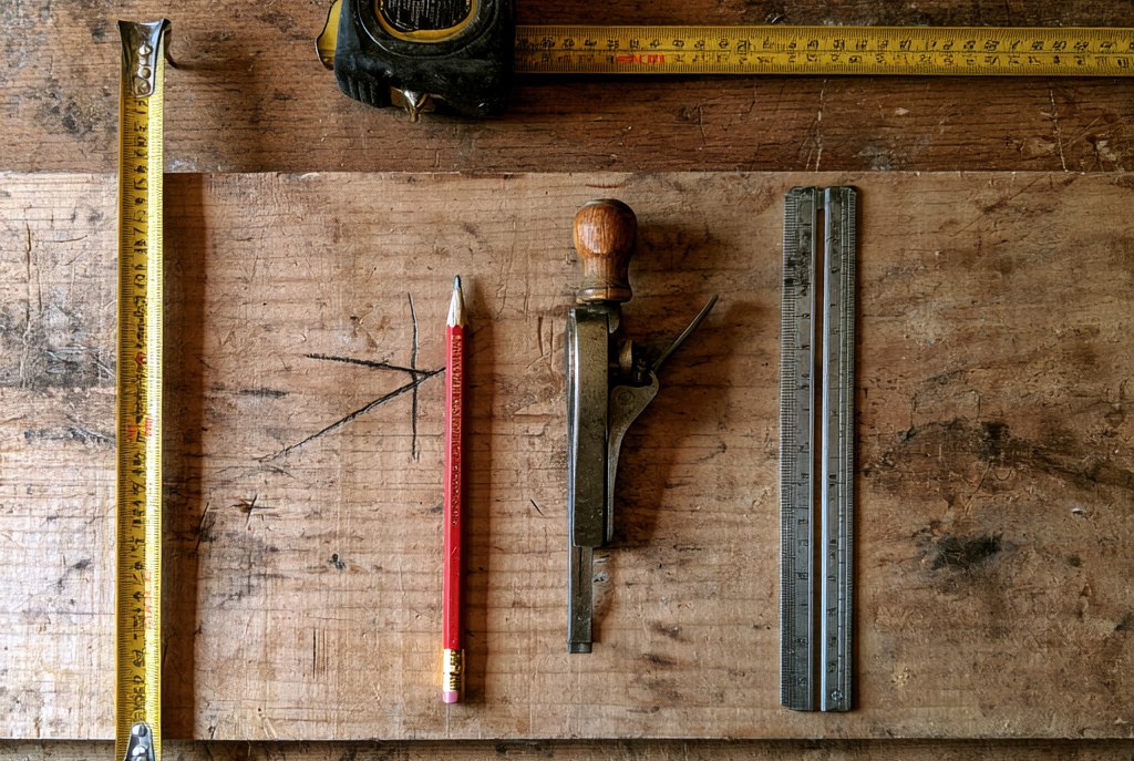 A workbench seen from above with a tape measure extended, a carpenter pencil, a marking gauge and a steel rule laid out on a board that carries pencil marks and a cross indicating the waste side
