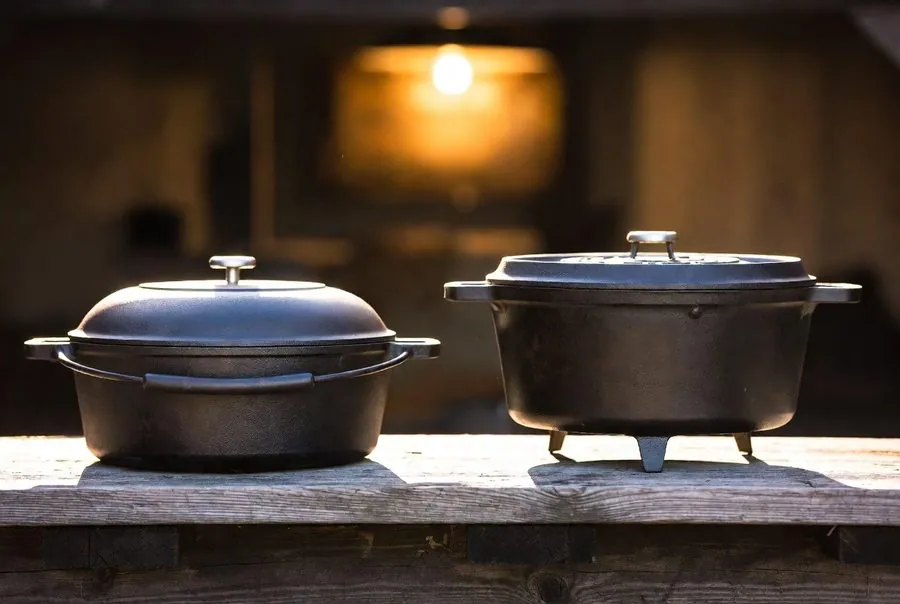 The two branches side by side on weathered wood: a flat-bottom kitchen Dutch oven and a three-legged camp Dutch oven built to stand over coals
