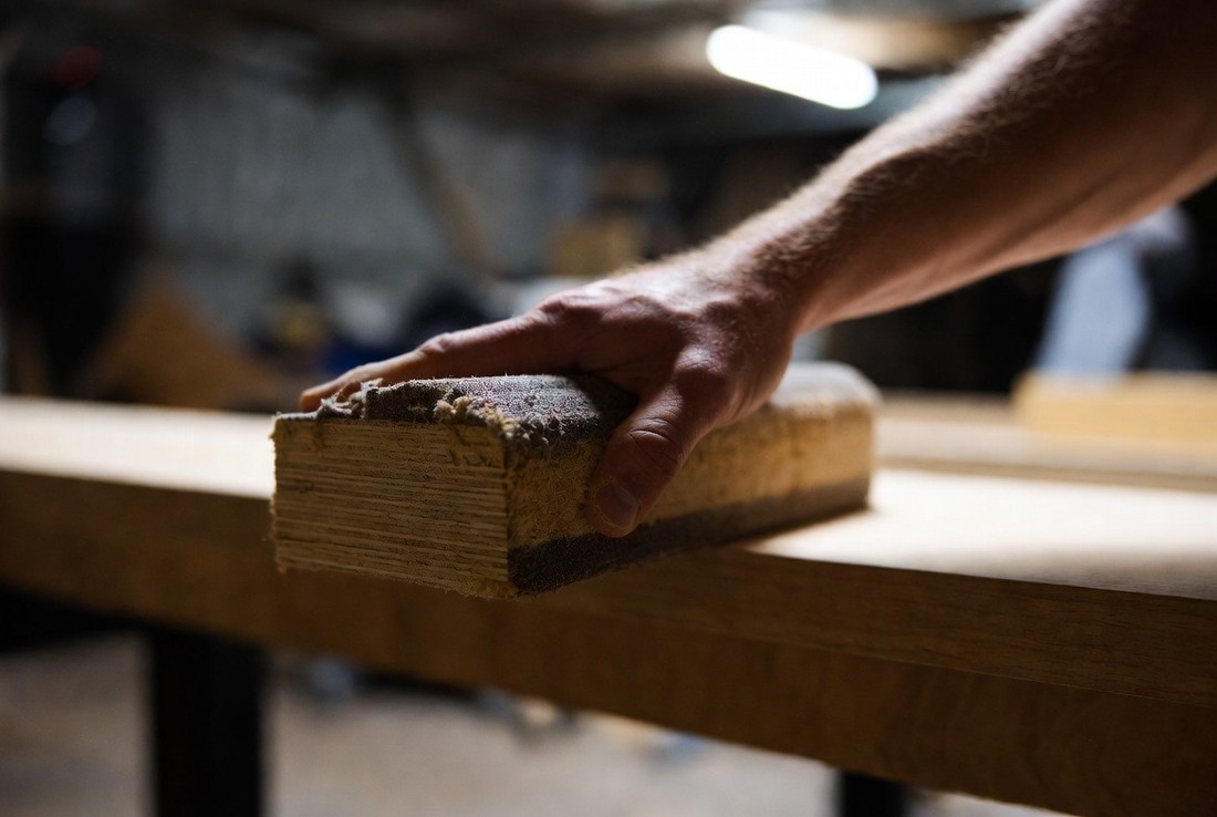 A hand pressing down hard on a worn, clogged sanding block on a workbench, the forearm leaning into it