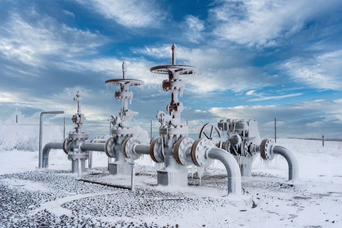 A natural gas pipeline valve station encased in ice and snow under a winter sky, valve wheels and pipework thick with frost
