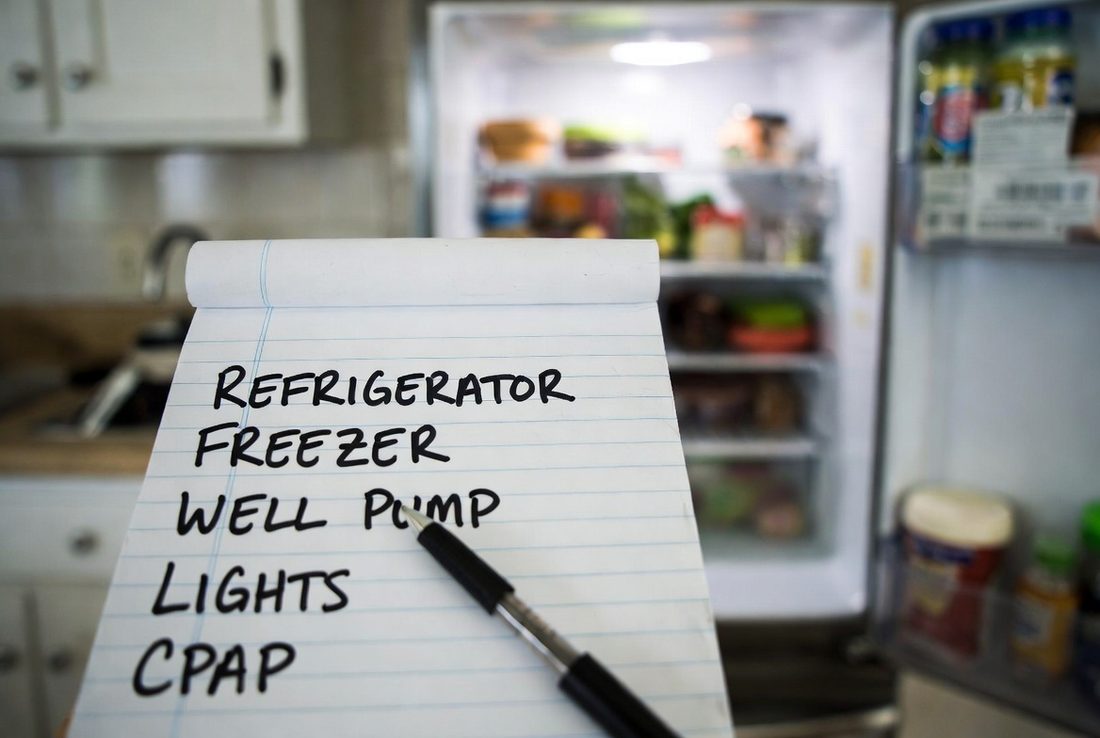 A yellow legal pad on a kitchen counter listing refrigerator, freezer, well pump, lights and CPAP in marker, a pen resting on it, an open refrigerator behind