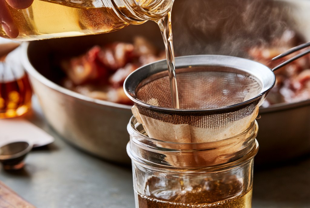 Warm liquid bacon fat being poured from a jar through a paper-lined fine mesh strainer into a clean glass jar, a skillet of cooked bacon behind, steam rising