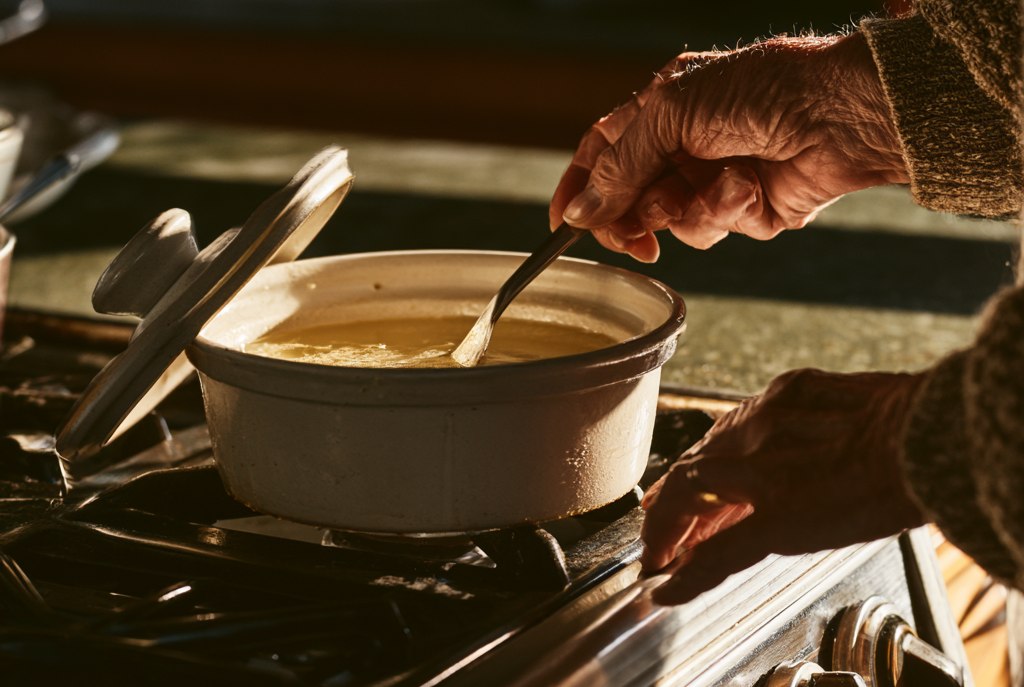 An older woman lifting a spoon from a cream ceramic crock of pale set bacon grease standing on a gas stove top, its lid propped against it, warm kitchen light