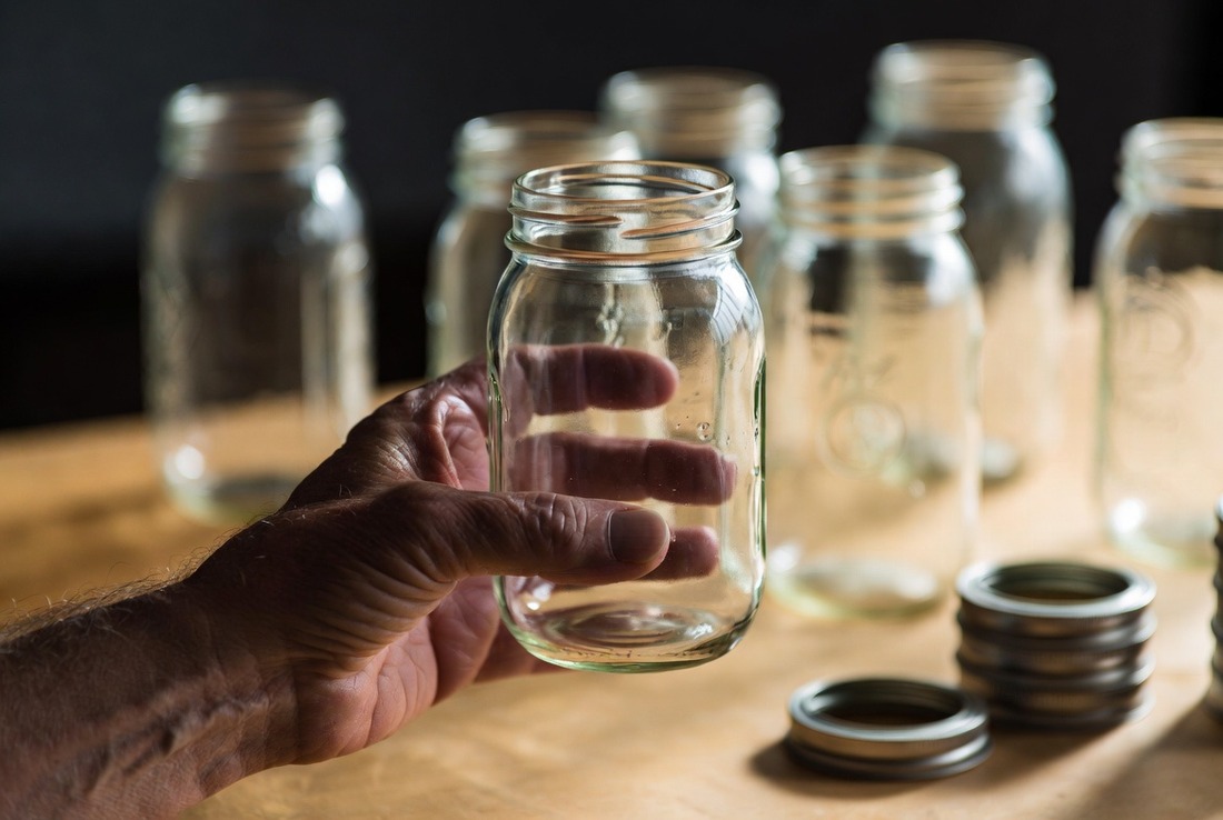 A weathered older hand holding an empty glass Mason jar on a wooden table, with more jars and a stack of metal rings behind it in soft focus