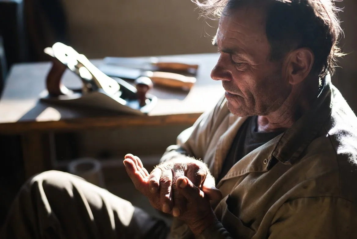 An older man in work clothes seated beside a bench of planes and chisels, looking down at his own open palm