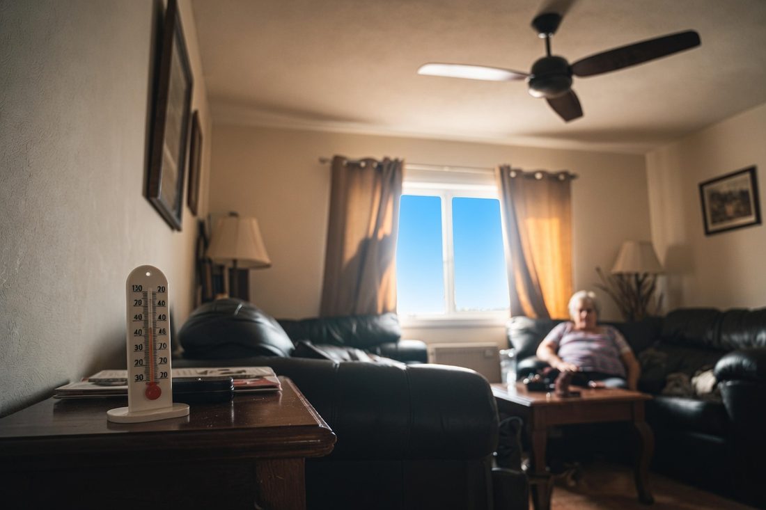 A dim living room on a bright day with the ceiling fan motionless, a thermometer standing on a side table in the foreground and an older woman sitting back on the sofa near a sunlit window