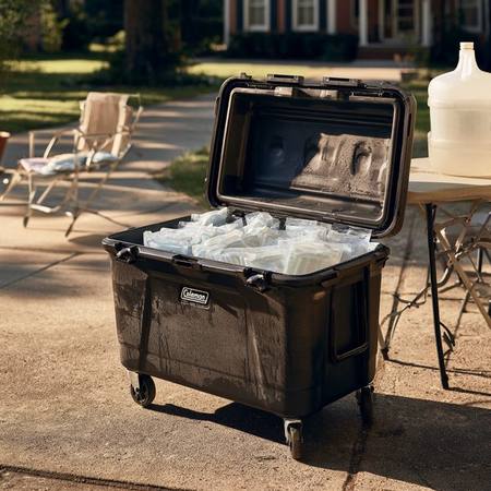 Wheeled Coleman cooler open on a sunny driveway filled with flat frozen water-filled sandwich bags as ice bricks, with a ten-gallon beverage jug on a folding table nearby