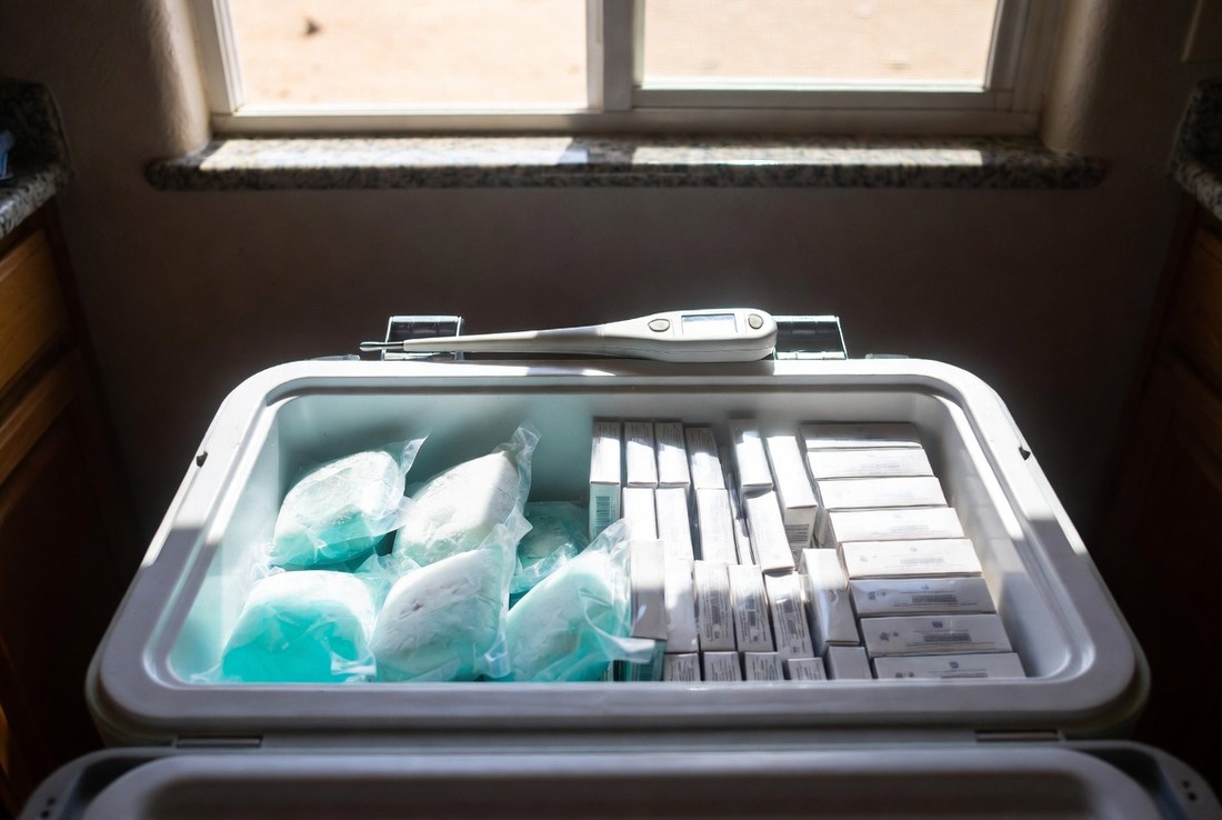 A cooler standing open on a kitchen counter packed with frozen gel packs on one side and rows of small closed medicine cartons on the other, a thermometer probe resting across the top