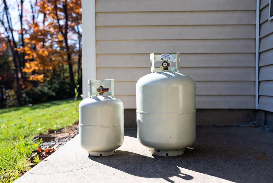 A twenty-pound propane cylinder standing beside a larger cylinder fitted with a gauge, both on a concrete pad against the siding of a house, autumn trees behind
