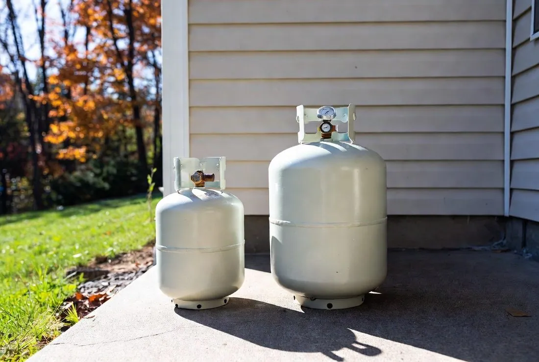 A twenty-pound propane cylinder standing beside a larger cylinder fitted with a gauge, both on a concrete pad against the siding of a house, autumn trees behind
