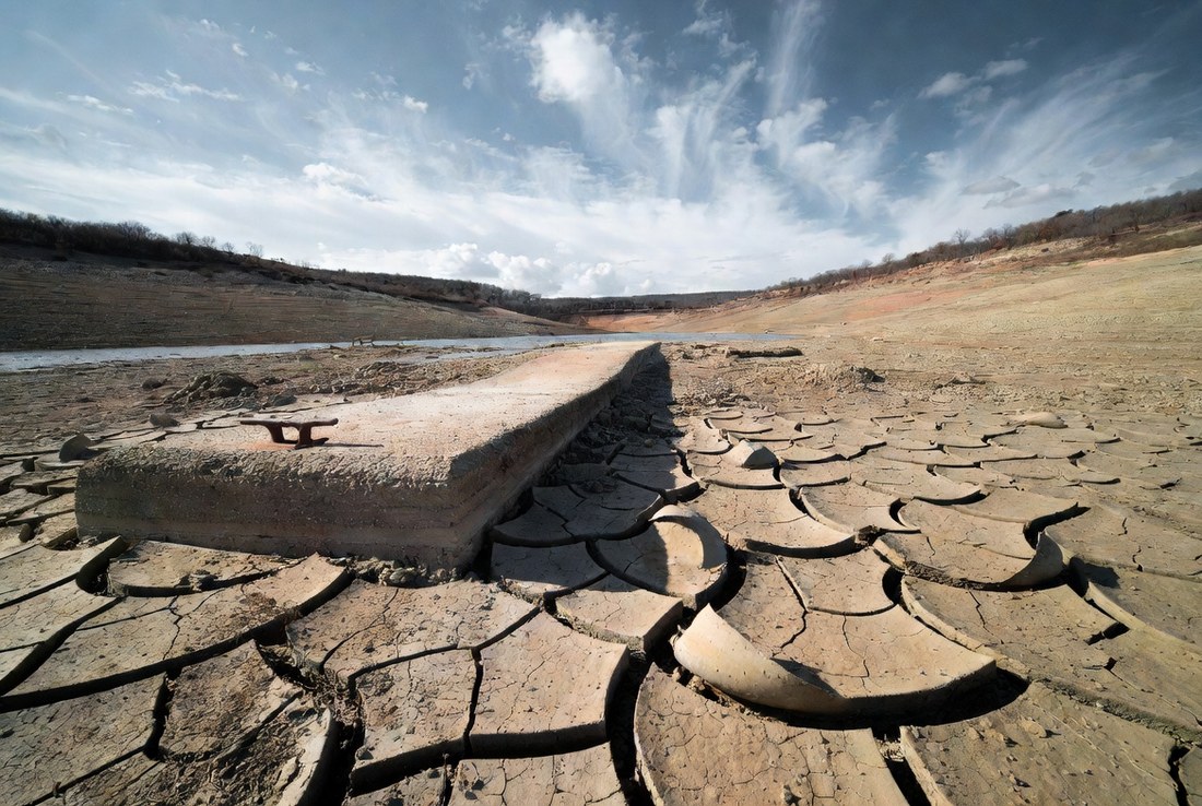 A dried cracked reservoir bed with a concrete boat ramp ending in open ground far short of the remaining water, a rusted mooring cleat still bolted in place
