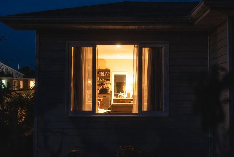 A lit living room seen from outside at night through a window with the curtains open, showing the bookcase, lamp, sofa and the doorway beyond