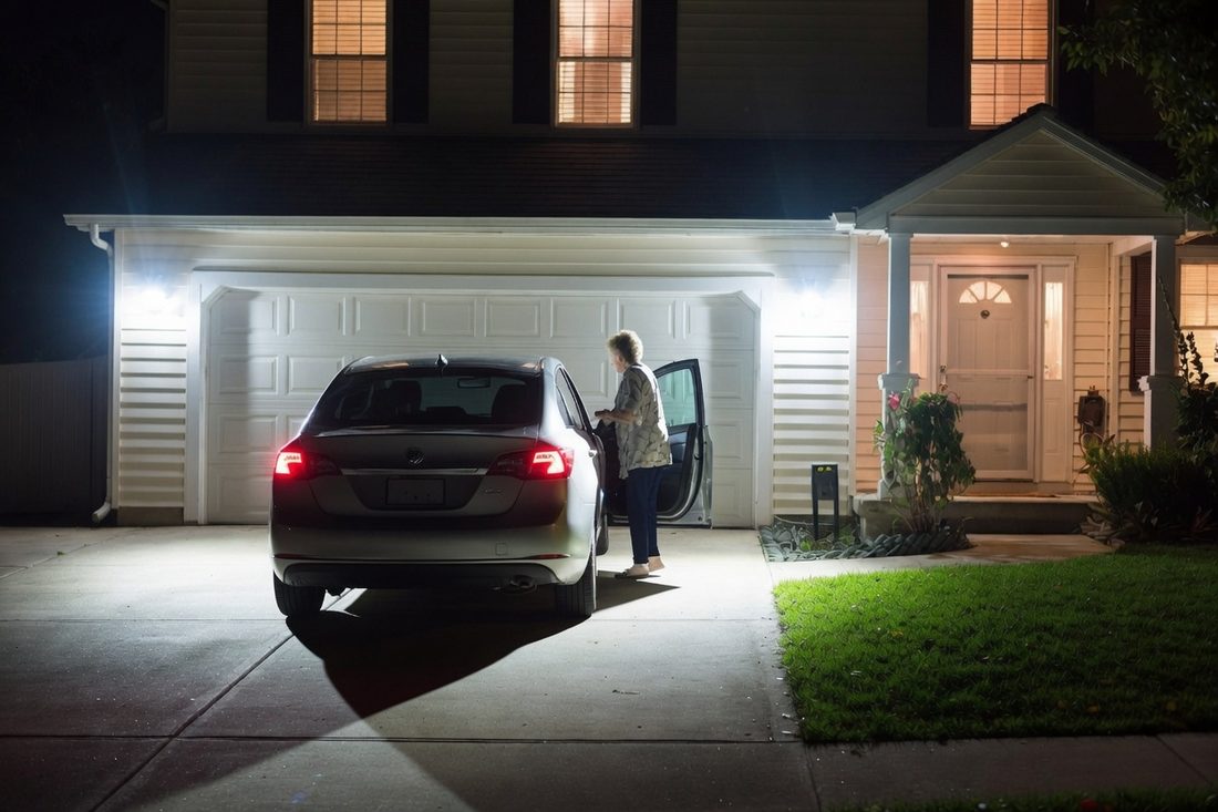 A car parked in a driveway at night with floodlights mounted above the garage door lighting the whole driveway and the ground beside the car, and an older woman getting out