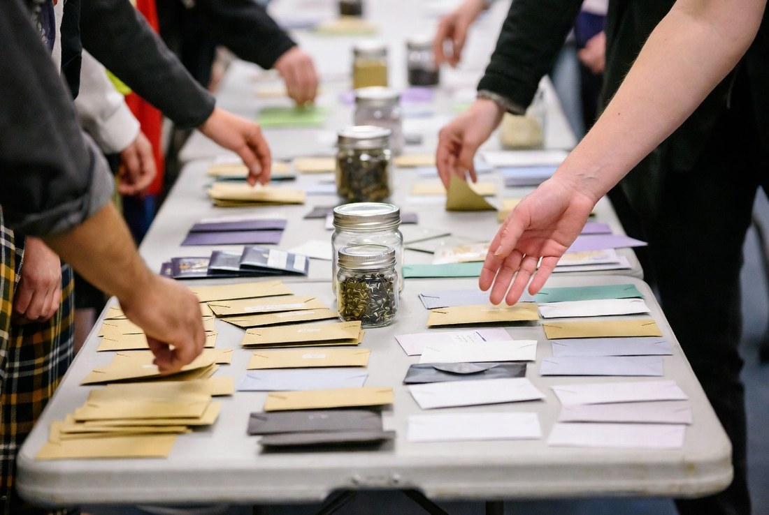 People leaning over a folding table at a seed swap, small paper envelopes and two jars of saved seed spread across it