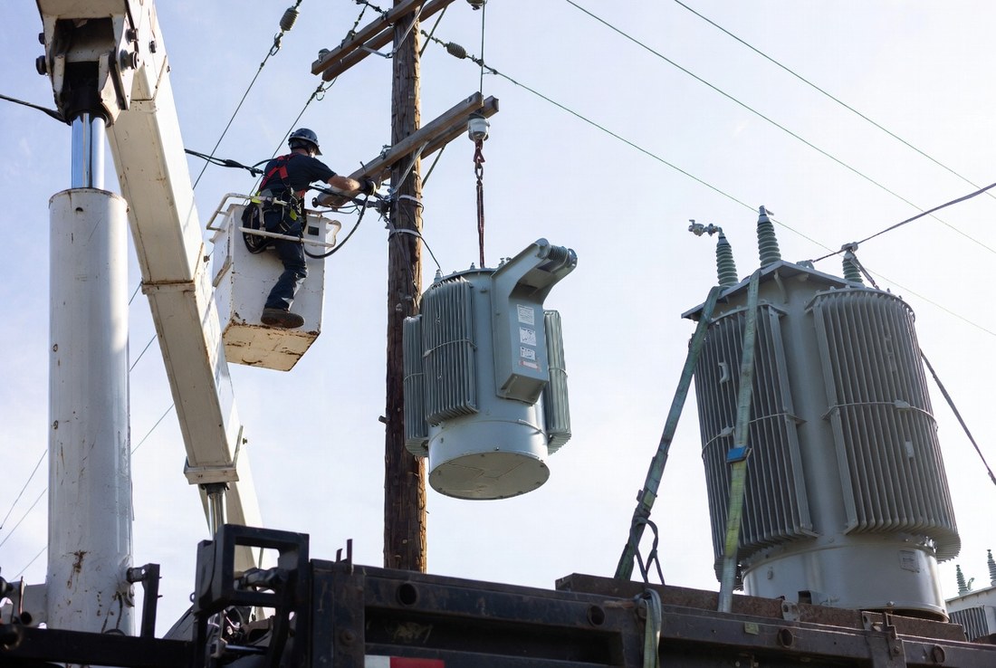 A pole-top distribution transformer suspended on a strap being lifted into position, seen from ground level against the sky