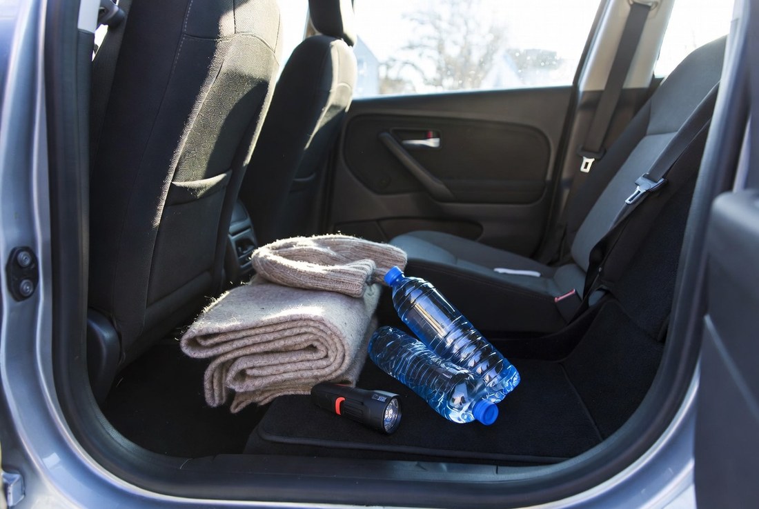 The rear footwell of a car holding a folded wool blanket, a knitted hat, two bottles of water and a small flashlight