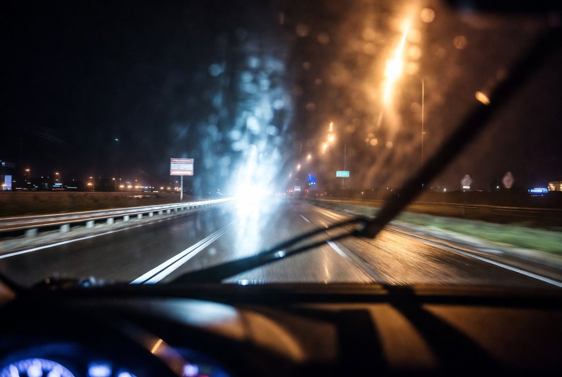 The view through a car windscreen at night on a wet road, oncoming headlights blooming across the wet glass with the lane markings barely readable