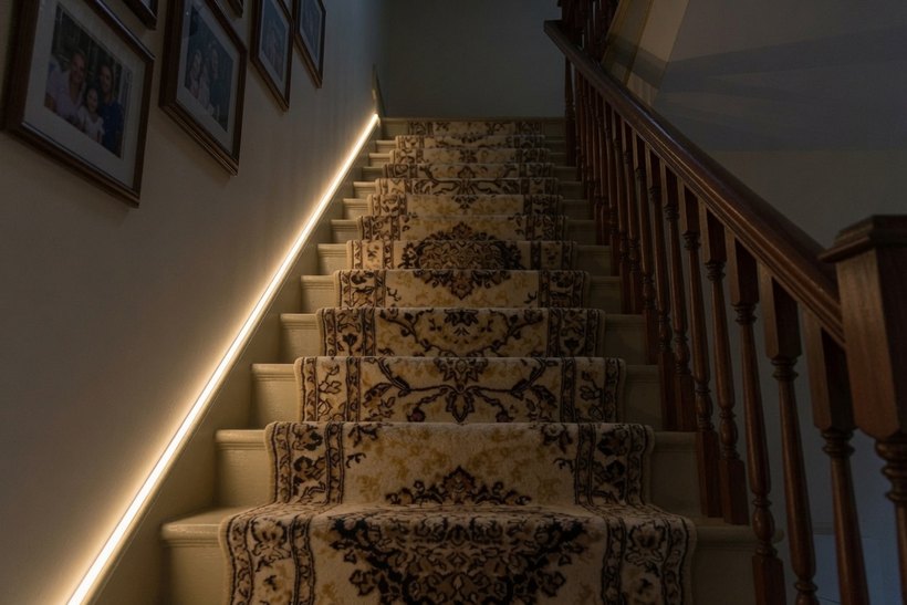 The same staircase at night with a continuous warm LED strip running up the wall at skirting height, the treads and the runner clearly visible